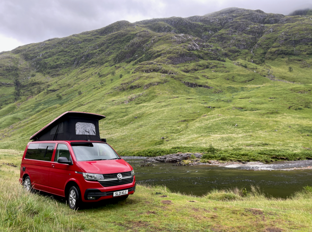 A red campervan parked in a scenic spot in front of a hill in Scotland